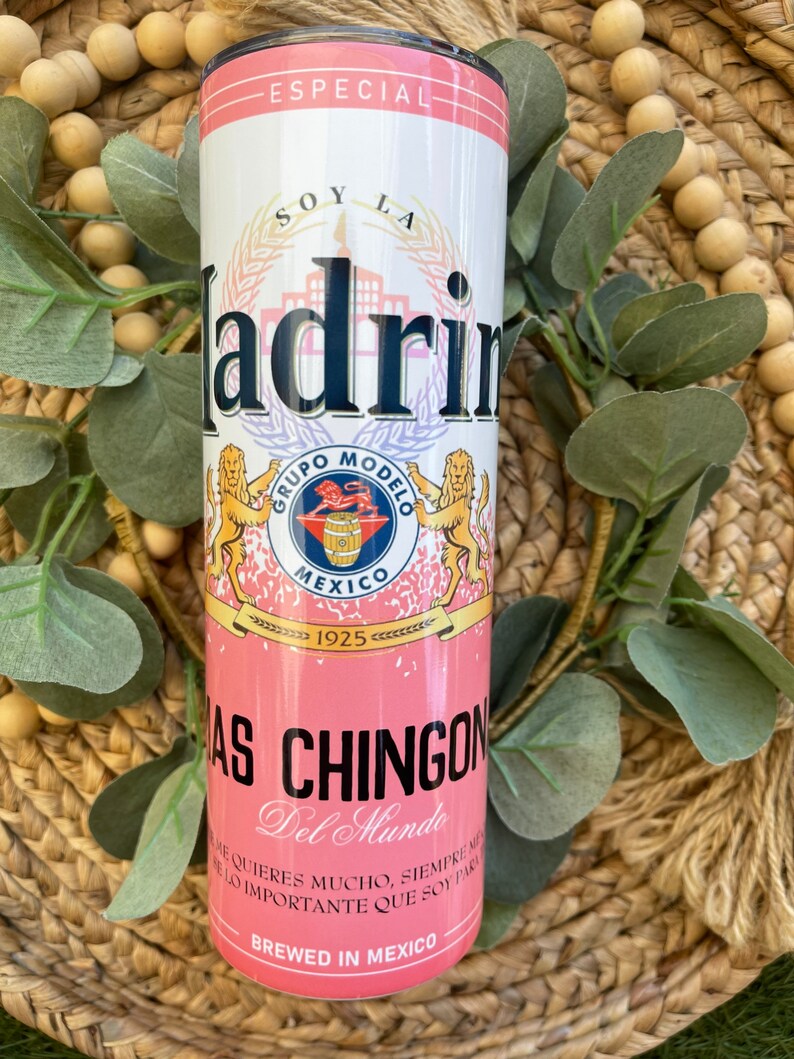 May include: Pink and white insulated tumbler with a graphic of a beer can. The can has the text "Soy La Madrin" and "Grupo Modelo" with a lion logo. The text "Especial" is at the top of the can and "Brewed in Mexico" is at the bottom.