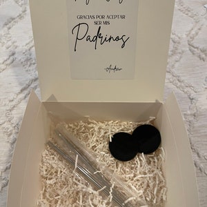May include: A white box with a white card inside. The card has black text that says "Gaby & Jorge" and "Gracias por aceptar ser mis Padrinos -Andrew". The box is filled with white shredded paper and contains two black lids and a set of silver straws.