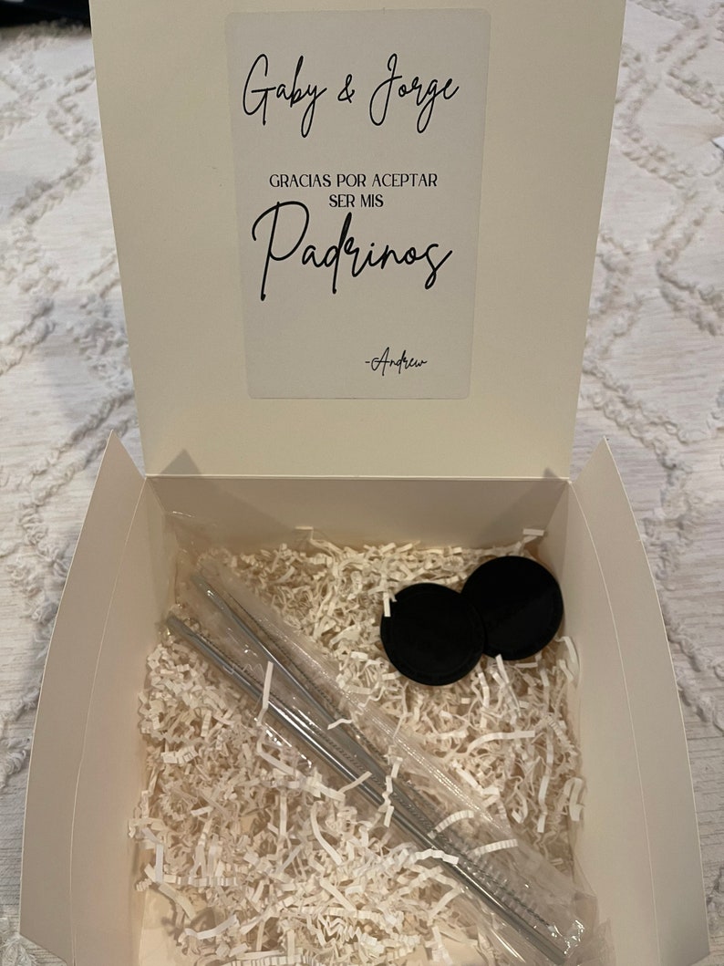 May include: A white box with a white card inside. The card has black text that says "Gaby & Jorge" and "Gracias por aceptar ser mis Padrinos -Andrew". The box is filled with white shredded paper and contains two black lids and a set of silver straws.
