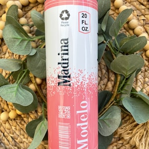 May include: Pink and white 20 fl oz tumbler with a design that resembles a beer can. The design features the words "Modelo" and "Madrina" with a barcode and a QR code. The tumbler has a black lid.