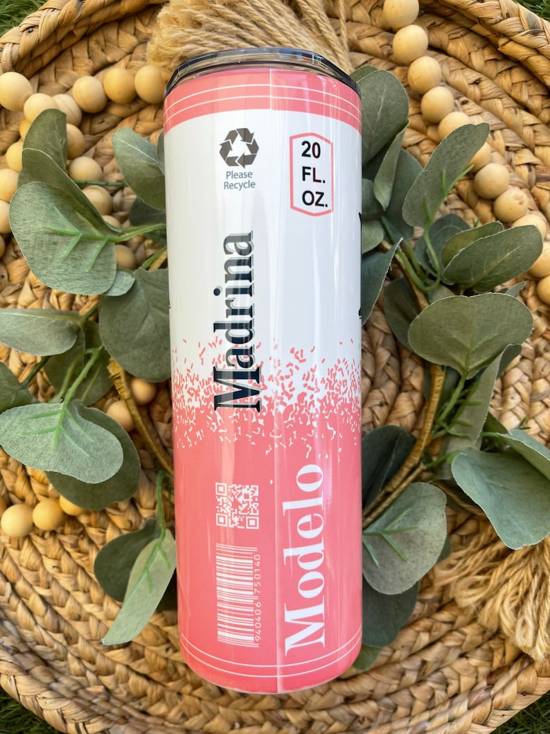 May include: Pink and white 20 fl oz tumbler with a design that resembles a beer can. The design features the words "Modelo" and "Madrina" with a barcode and a QR code. The tumbler has a black lid.