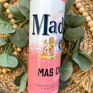 May include: A pink and white insulated tumbler with a black lid. The tumbler features a graphic of a lion and the text "Mad" and "Mas Chido" in black. The text "Porque me quieres mas y se lo importante" is printed in black at the bottom of the tumbler.