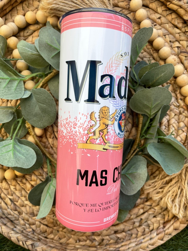 May include: A pink and white insulated tumbler with a black lid. The tumbler features a graphic of a lion and the text "Mad" and "Mas Chido" in black. The text "Porque me quieres mas y se lo importante" is printed in black at the bottom of the tumbler.
