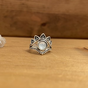 May include: A silver-toned ring with a central, round, light-colored stone. The ring features a detailed design with a floral or sunburst-like pattern above the stone and a beaded edge. The ring is set against a wooden background.
