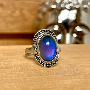 Può includere: Un anello d'argento con una grande pietra ovale di umore che cambia colore. La pietra è incastonata in una ghiera d'argento con un bordo decorativo.