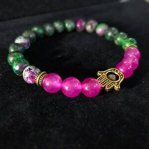 May include: A pink and green gemstone bracelet with a gold hamsa charm. The bracelet is made of round beads and is strung on elastic.