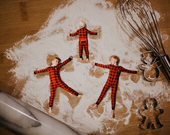 May include: Three children in red and black plaid pajamas make snow angels in a pile of white flour on a wooden surface. A rolling pin and a whisk are also visible in the image.