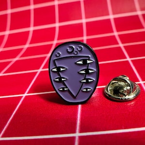 May include: Purple enamel pin with a black outline. The pin features a triangle with six eyes and three crescent moons. The pin is on a red grid background.