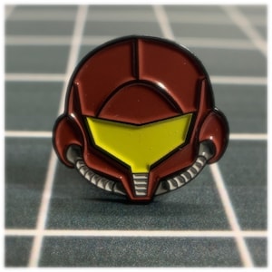 May include: A red and yellow enamel pin featuring a stylized depiction of a video game character's helmet.