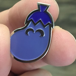 Puede incluir: Un pin de esmalte de berenjena morado con una parte superior azul y una cara sonriente.