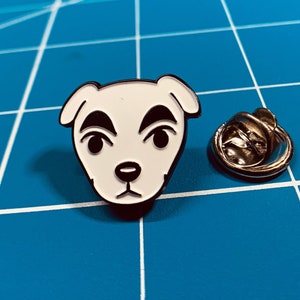 May include: White enamel pin of a dog with black eyes and eyebrows. The pin has a silver metal backing.