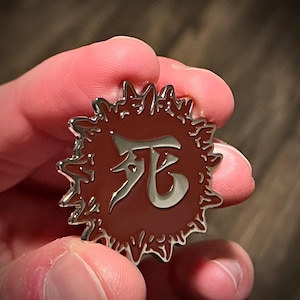 Könnte beinhalten: Eine silberne Anstecknadel mit rotem Hintergrund und einem weißen Kanji-Zeichen, das im Zentrum "Tod" bedeutet. Die Nadel ist von einem gezackten, stacheligen Rand umgeben.