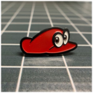 May include: A red and black enamel pin shaped like a hat with two large white eyes.