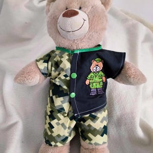May include: A plush tan teddy bear wearing a two-toned outfit. The left side features a green, tan, and black digital camouflage pattern, while the right side is solid black with an embroidered bear graphic wearing a green uniform and two green buttons.