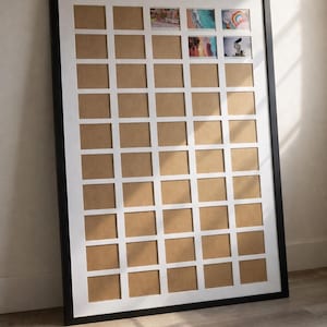 May include: A large black framed photo collage with a white grid of 42 square openings. Some openings contain colorful photos. The frame is leaning against a wall. The overall theme is home decor.