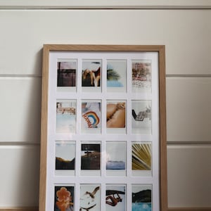 May include: A wooden frame with a white mat holding 16 Polaroid photos. The photos are arranged in a grid pattern and feature a variety of subjects, including landscapes, people, and objects. The frame is on a wooden shelf.