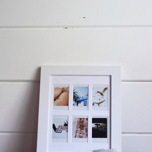 May include: A white picture frame with six Polaroid photos inside. The photos show a variety of scenes, including a woman in a bikini, a motel sign, a street scene, and a car driving down a road. The frame is sitting on a black surface with a clear quartz crystal in front of it.
