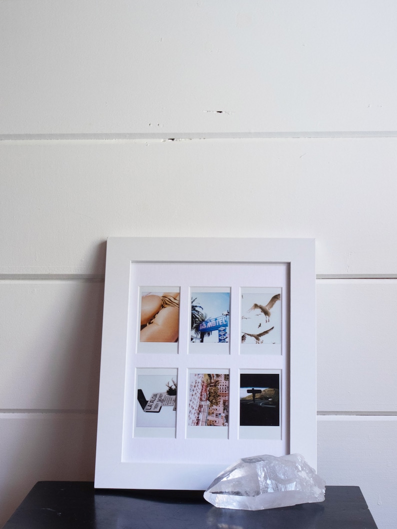 May include: A white picture frame with six Polaroid photos inside. The photos show a variety of scenes, including a woman in a bikini, a motel sign, a street scene, and a car driving down a road. The frame is sitting on a black surface with a clear quartz crystal in front of it.