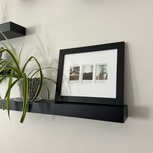 May include: A black floating shelf with a black picture frame on top. The picture frame has three black and white photos inside. A green plant in a white pot is on the shelf.