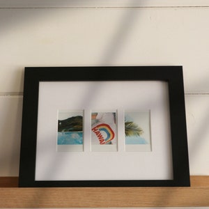 Puede incluir: Un marco de fotos negro con un paspartú blanco y tres fotos Polaroid en su interior. Las fotos muestran una escena de playa, una persona que lleva una camiseta que dice "HAWAII" con un arcoíris, y una palmera.
