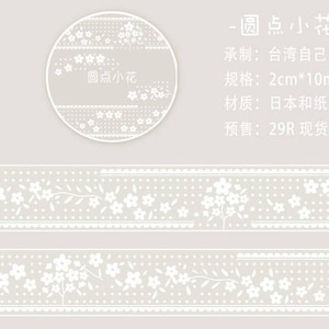 Pode incluir: Fita washi branca com um padrão repetitivo de pequenas flores brancas e pontos sobre um fundo cinza claro. A fita é etiquetada "圆点小花" que se traduz como "flor pequena com pontos circulares" em inglês. A fita mede 2 cm de largura e 10 m de comprimento.