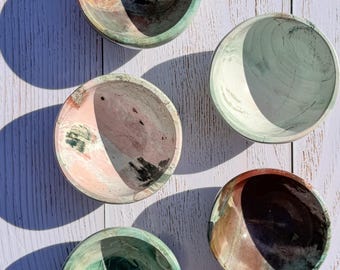 May include: Five small, round bowls with a marbled appearance. The bowls feature a mix of colors, including green, pink, brown, and black. The bowls are arranged on a light-colored wooden surface, with shadows cast across them.