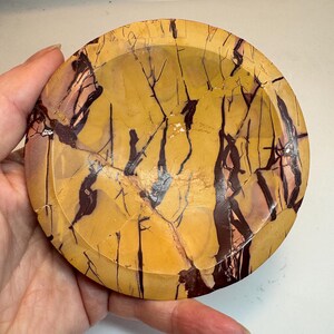 Puede incluir: Un plato redondo de piedra pulida con un patrón único. El plato presenta una base de tonos amarillos y beige, con marcas oscuras y negras en forma de vetas. El plato está sostenido en una mano.