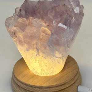 May include: A lit amethyst crystal lamp. The crystal cluster is a light purple colour and sits atop a round, wooden base. The lamp emits a warm, soft glow. A white power cord is plugged into the base.