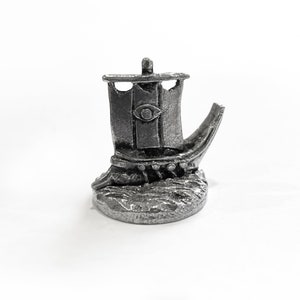 Puede incluir: Una miniatura de plata de estaño de un barco con una sola vela y un diseño de ojo en la vela. El barco está sentado sobre una base que parece agua.