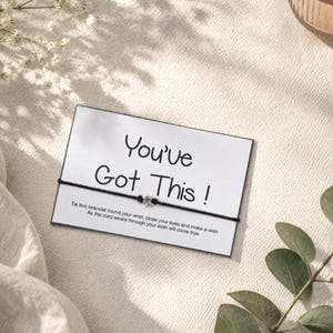 May include: A card with the text "You've Got This!" is displayed with a black string bracelet. The card is on a white surface, with a lit candle in a glass holder and sprigs of greenery. Soft shadows add depth.