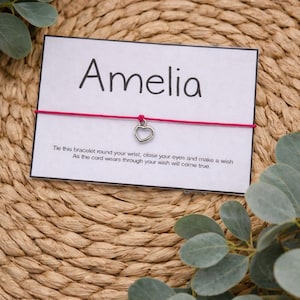 May include: A pink string bracelet with a silver heart charm, presented on a white card with the name "Amelia". The card also includes text about making a wish. The background is a woven, natural-toned mat with green foliage.