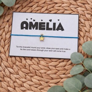May include: A blue string bracelet with a gold star charm, presented on a card that reads "AMELIA" in bold letters. The card also includes a message about making a wish. The background is a woven texture with green leaves.