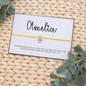 May include: A yellow cord bracelet with a silver star charm, presented on a white card with the name "Amelia" in cursive. The card includes a message about making a wish. The background is a woven mat with eucalyptus leaves.