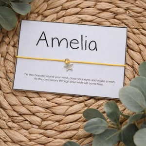 May include: A personalized wish bracelet with the name "Amelia" printed on a white card. The card features a yellow cord with a silver star charm. The text on the card encourages the wearer to make a wish.