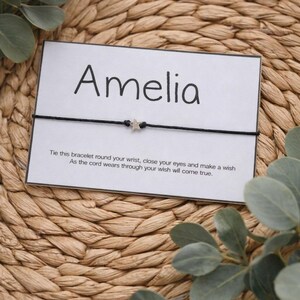 May include: A black string bracelet with a small silver star charm is displayed on a white card. The card is printed with the name "Amelia" and a wish-making message. The background is a woven texture with green leaves.