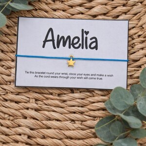 May include: A blue string bracelet with a gold star charm, presented on a white card with the name "Amelia" in black script. The card includes a wish-making message. The background is a woven wicker texture.
