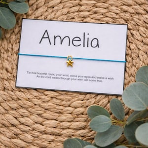 May include: A teal string bracelet with a gold star charm, presented on a white card with the name "Amelia" printed on it. The card also includes a wish-making message. The background is a woven texture with eucalyptus leaves.