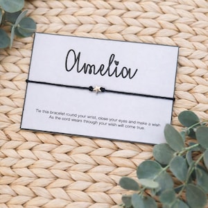 Op de afbeelding: Een zwart koordarmbandje met een kleine sterbedel, gepresenteerd op een kaart met de tekst "Amelia". De kaart bevat ook de tekst: "Bind deze armband om je pols, sluit je ogen en doe een wens. Naarmate het koord slijt, komt je wens uit."