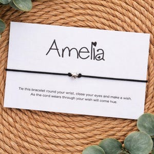 Op de afbeelding: Een witte kaart met de naam "Amelia" in zwart schrift, een zwart koordarmbandje met een sterbedel en de tekst "Bind deze armband om je pols, sluit je ogen en doe een wens."