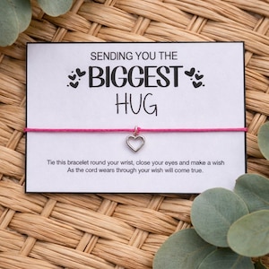 May include: A card with the message "SENDING YOU THE BIGGEST HUG" and a pink cord bracelet with a heart pendant. The card has instructions to tie the bracelet and make a wish. The background is woven.