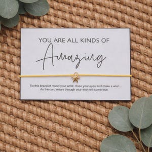 May include: A yellow string bracelet with a gold star charm is displayed on a white card with the words "YOU ARE ALL KINDS OF Amazing". The card also includes instructions. The background is a woven, tan surface.