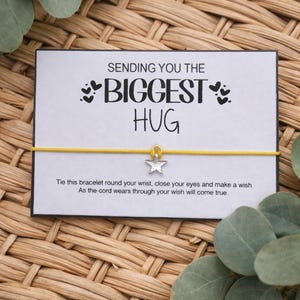 May include: A white card with the message "SENDING YOU THE BIGGEST HUG" and a yellow bracelet with a star charm. The card includes instructions to tie the bracelet and make a wish. The background is a woven texture with green leaves.