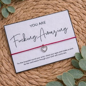 May include: A red string bracelet with a silver heart charm, presented on a white card. The card reads "YOU ARE Fucking Amazing" in cursive script, with additional text below. The background is a woven texture.