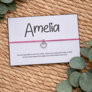 May include: A pink cord bracelet with a silver heart charm, presented on a card with the name "Amelia" in black script. The card includes a message about making a wish. The background is a woven texture.