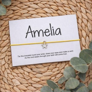 May include: A white card with the name "Amelia" in black script, a yellow string, and a silver star charm. The card includes text that reads, "Tie this bracelet round your wrist, close your eyes and make a wish. As the cord wears through your wish will come true."