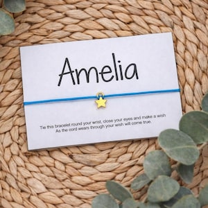 May include: A card with the name "Amelia" printed in black, with a blue string bracelet and a gold star charm. The card has text about making a wish. The background is a woven texture.