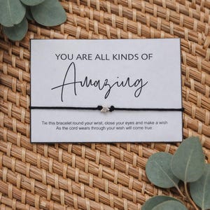 May include: A black cord bracelet with a silver star charm, displayed on a card with the words "YOU ARE ALL KINDS OF Amazing". The card rests on a woven wicker surface, with eucalyptus leaves.