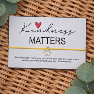 May include: A card with the words "Kindness Matters" and a yellow string bracelet with a star charm. The card has a red heart and text that reads, "Tie this bracelet round your wrist, close your eyes and make a wish. As the cord wears through your wish will come true."