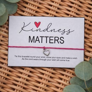 May include: A white card with the words "Kindness Matters" and a red heart graphic. A pink string bracelet with a silver heart charm is attached. The card is on a woven background with green leaves.