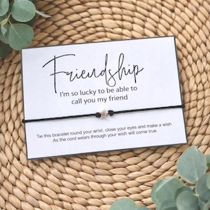 May include: A friendship wish bracelet on a white card with the word "Friendship" in script. The card also includes the text "I'm so lucky to be able to call you my friend." and instructions. The bracelet has a black cord and a star charm.